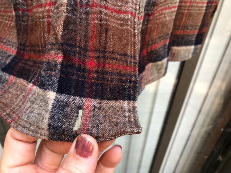 May include: Close-up of a brown, red, and navy blue plaid fabric. The fabric has a textured appearance with visible woven threads. The hem of the garment is visible, with a small opening. The overall color palette is warm and rustic.