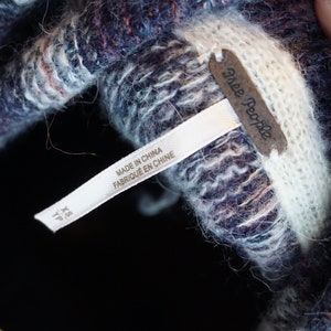 May include: Close-up of a textured knit garment in shades of blue, purple, and white. The garment has a tag that reads "Free People" and "Made in China". The size is XS/TP.