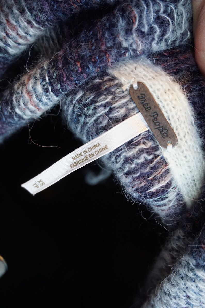 May include: Close-up of a textured knit garment in shades of blue, purple, and white. The garment has a tag that reads "Free People" and "Made in China". The size is XS/TP.