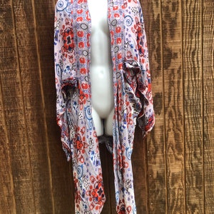 May include: A lightweight, open-front kimono-style cardigan with a floral pattern in red, blue, and white on a light purple background. The cardigan is displayed on a mannequin against a wooden backdrop.