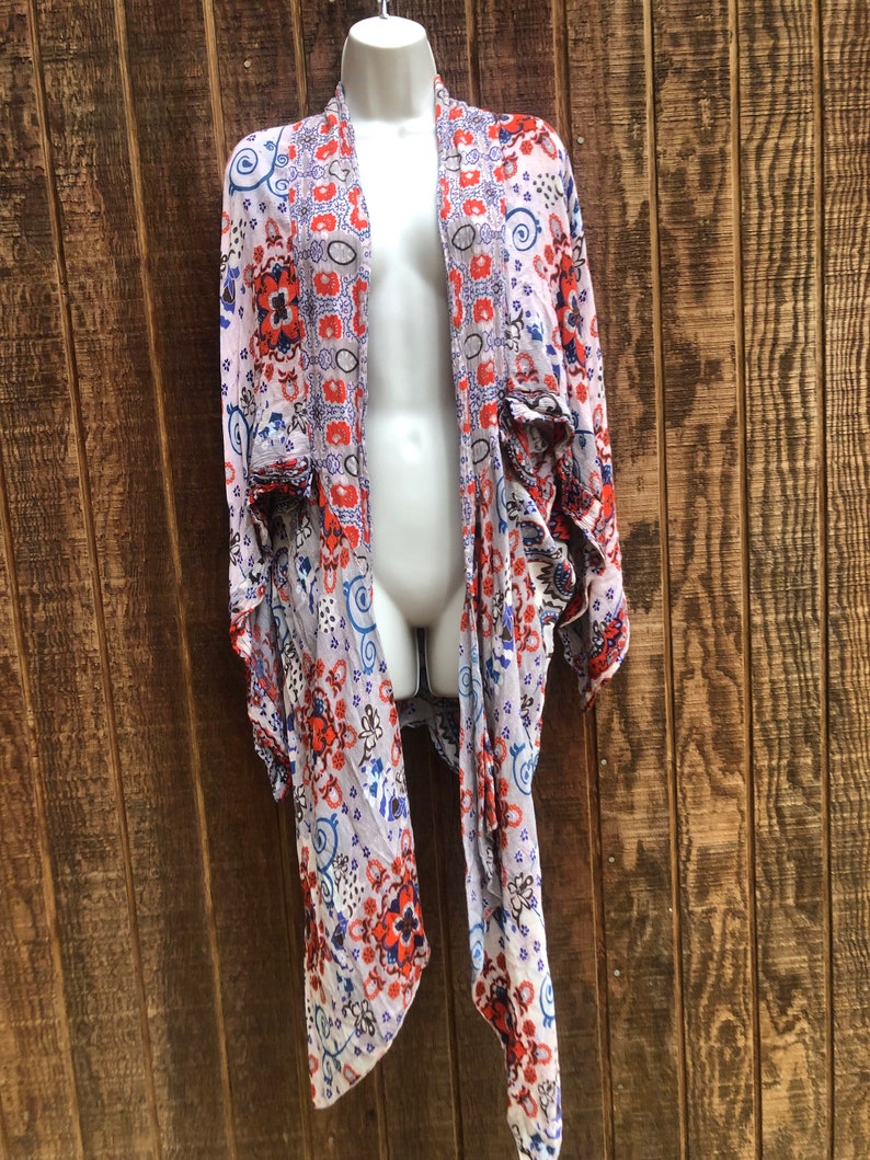 May include: A lightweight, open-front kimono-style cardigan with a floral pattern in red, blue, and white on a light purple background. The cardigan is displayed on a mannequin against a wooden backdrop.