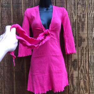 May include: A bright pink dress with a V-neckline and long sleeves. The dress features a tie detail at the waist and a ruffled hem. The dress is displayed on a black mannequin against a wooden background.