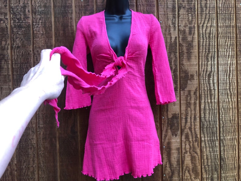 May include: A bright pink dress with a V-neckline and long sleeves. The dress features a tie detail at the waist and a ruffled hem. The dress is displayed on a black mannequin against a wooden background.