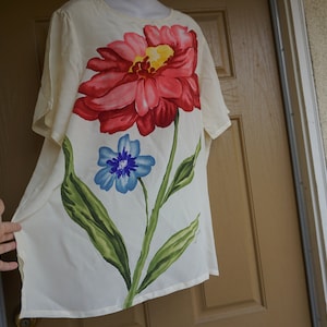 May include: Cream-colored, short-sleeved top featuring a large floral design. The artwork includes a large red and pink flower with a yellow center, a smaller blue flower, and green leaves. The garment is displayed on a mannequin.