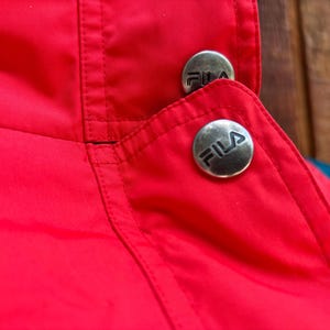 May include: Close-up of a red jacket collar with two silver buttons. Each button has the FILA logo in black. The jacket has a blue and teal accent.