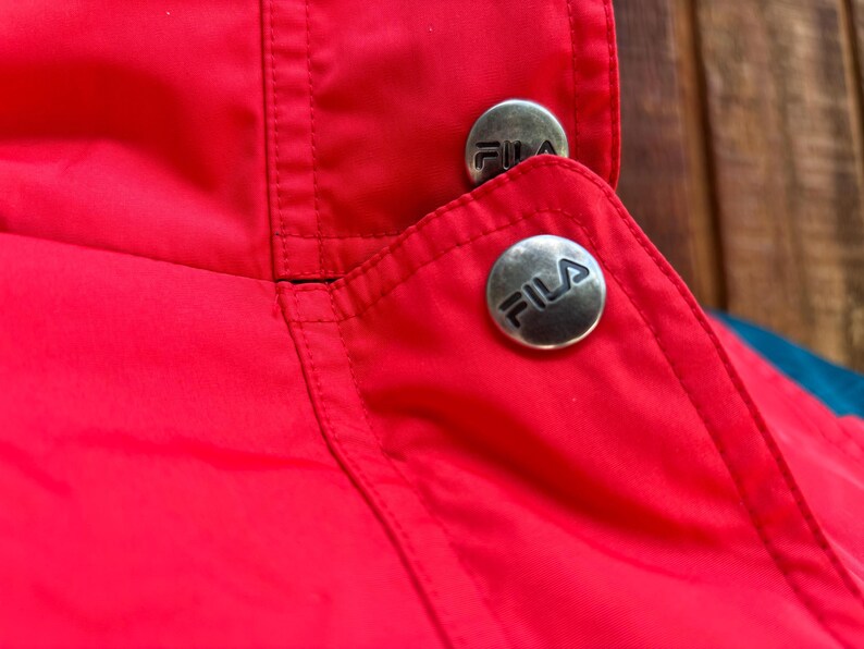 May include: Close-up of a red jacket collar with two silver buttons. Each button has the FILA logo in black. The jacket has a blue and teal accent.