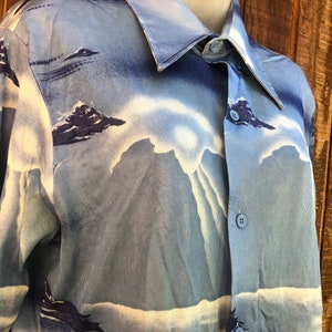 May include: Light blue button-down shirt with a mountain and cloud design. The shirt features a collared neckline and long sleeves. The mountain design is in shades of blue and white, creating a scenic landscape.