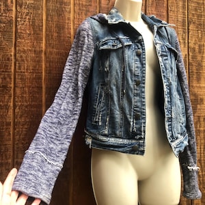 May include: A blue denim jacket with a hood and gray knit sleeves. The jacket has a button-up front, pockets, and distressed details. The sleeves have a textured appearance and the cuffs are trimmed with white stitching.