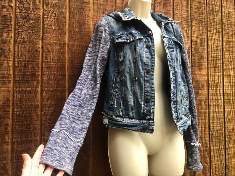 May include: A blue denim jacket with a hood and gray knit sleeves. The jacket has a button-up front, pockets, and distressed details. The sleeves have a textured appearance and the cuffs are trimmed with white stitching.