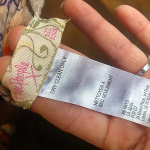 May include: A tag from the brand "Free People X" with the text "DRY CLEAN ONLY" and "NETTOYER &Agrave; SEC SEULEMENT". The tag is attached to a patterned fabric with orange, blue, and white floral designs.