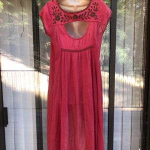 May include: A bright pink dress with short sleeves and a keyhole neckline. The dress features brown embroidered floral detailing around the neckline and a similar design at the waist. The dress is hanging on a white mannequin.