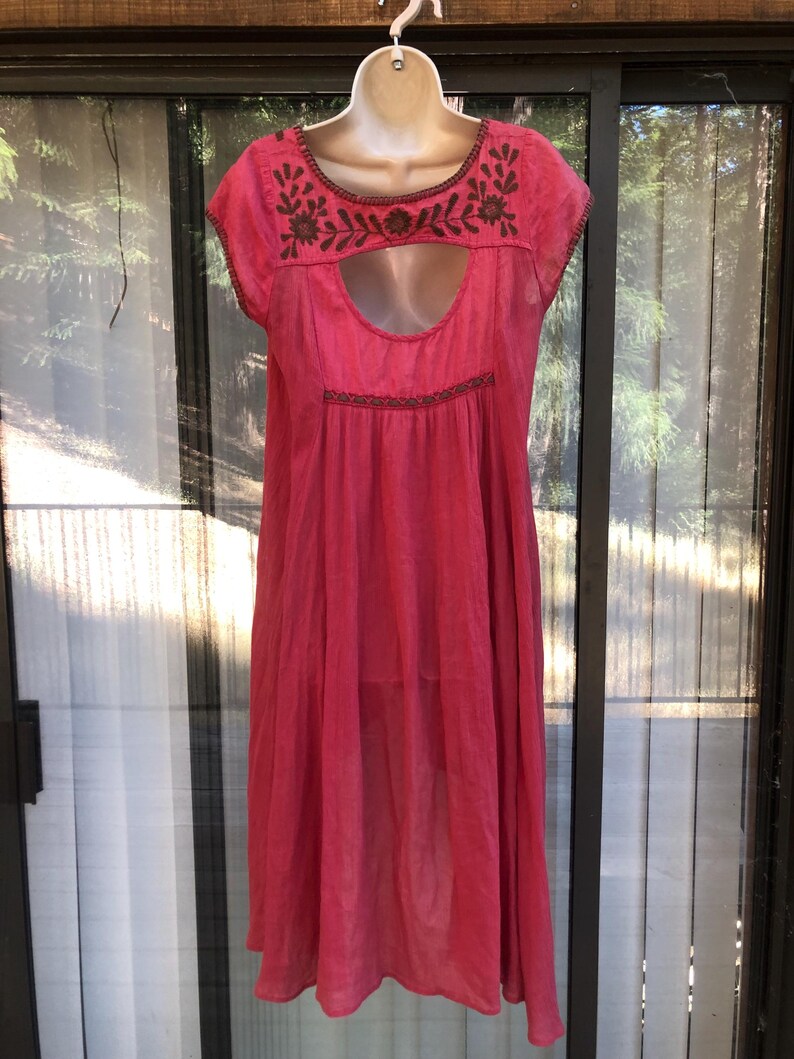 May include: A bright pink dress with short sleeves and a keyhole neckline. The dress features brown embroidered floral detailing around the neckline and a similar design at the waist. The dress is hanging on a white mannequin.
