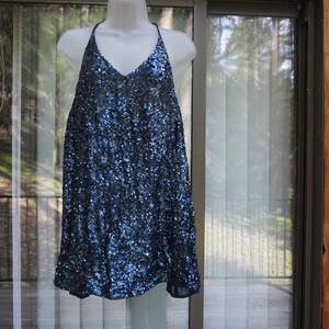 K&ouml;nnte beinhalten: Ein marineblaues Paillettenkleid mit Neckholder-Ausschnitt und V-Ausschnitt. Das Kleid wird auf einer wei&szlig;en Schaufensterpuppe pr&auml;sentiert. Das Kleid ist knielang und hat eine lockere, flie&szlig;ende Silhouette. Der Hintergrund umfasst ein Fenster und eine Holzwand.