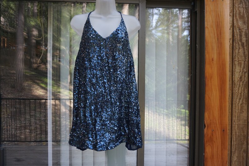 K&ouml;nnte beinhalten: Ein marineblaues Paillettenkleid mit Neckholder-Ausschnitt und V-Ausschnitt. Das Kleid wird auf einer wei&szlig;en Schaufensterpuppe pr&auml;sentiert. Das Kleid ist knielang und hat eine lockere, flie&szlig;ende Silhouette. Der Hintergrund umfasst ein Fenster und eine Holzwand.