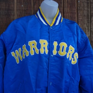May include: A blue satin jacket with the word "WARRIORS" in yellow lettering across the chest. The jacket has a yellow, white, and blue striped collar and cuffs. The jacket is buttoned up and appears to be vintage.