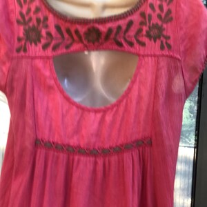 May include: A bright pink, short-sleeved top with brown embroidered floral and leaf designs around the neckline and a keyhole cutout. The top has a gathered waist and a simple, flowing design.