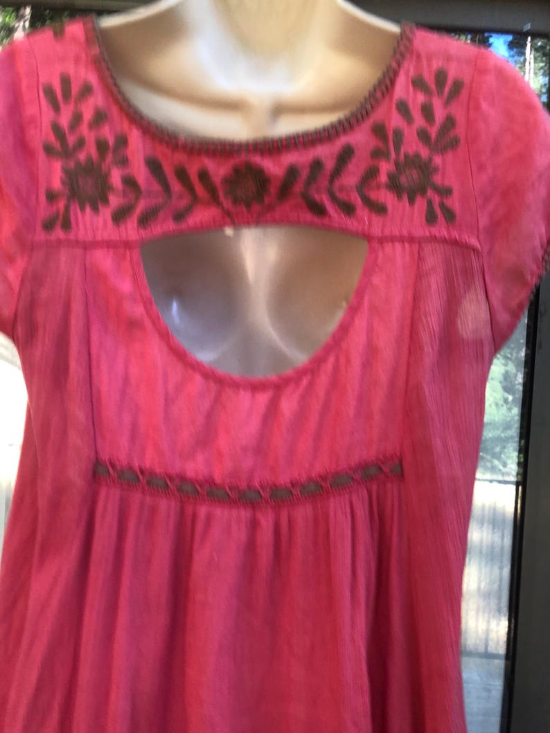 May include: A bright pink, short-sleeved top with brown embroidered floral and leaf designs around the neckline and a keyhole cutout. The top has a gathered waist and a simple, flowing design.