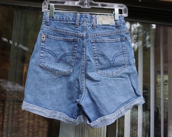 Puede incluir: Pantalones cortos vaqueros azul claro con dobladillos enrollados, colgados de una percha de plástico transparente. Los pantalones cortos tienen dos bolsillos traseros con un diseño curvo distintivo y un pequeño logotipo en el bolsillo derecho. Estilo clásico.