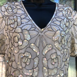 May include: A light-colored, short-sleeved top with a V-neck, adorned with floral patterns. The design features sequins, beads, and dark trim, creating a shimmering effect. The garment is displayed on a black mannequin.