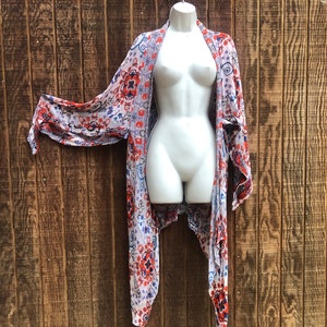 May include: A lightweight, open-front kimono-style cover-up with a colorful floral pattern in red, blue, and white. The fabric drapes over a white mannequin, showcasing the garment's flowing design. The background is a rustic wooden wall.