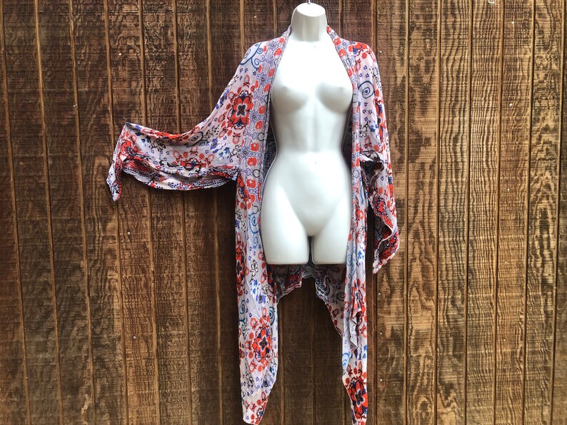 May include: A lightweight, open-front kimono-style cover-up with a colorful floral pattern in red, blue, and white. The fabric drapes over a white mannequin, showcasing the garment's flowing design. The background is a rustic wooden wall.