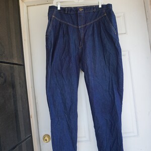 May include: Dark blue denim trousers with a high waist and pleated front. The trousers are hanging on a white hanger against a white door. The trousers have a button closure and pockets.