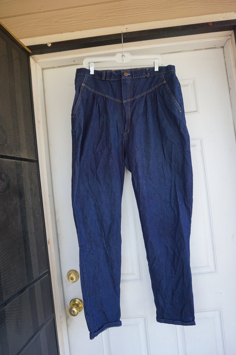 May include: Dark blue denim trousers with a high waist and pleated front. The trousers are hanging on a white hanger against a white door. The trousers have a button closure and pockets.