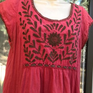 May include: A pink embroidered dress with short sleeves. The dress features brown floral embroidery across the chest and neckline. The fabric has a textured appearance.