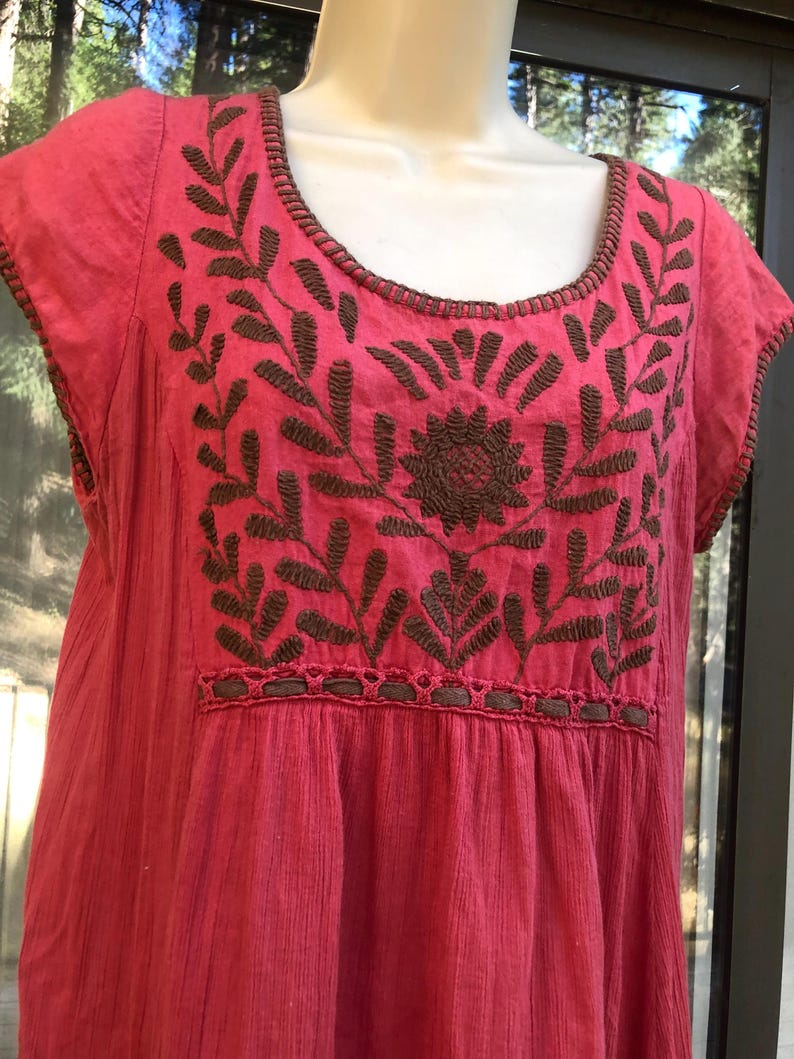 May include: A pink embroidered dress with short sleeves. The dress features brown floral embroidery across the chest and neckline. The fabric has a textured appearance.