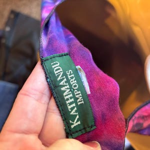 May include: A tie-dye garment featuring vibrant shades of purple, pink, and blue. A rectangular green fabric label is stitched onto the inner neckline, displaying the white text KATHMANDU IMPORTS in capital letters with a smaller tag underneath.