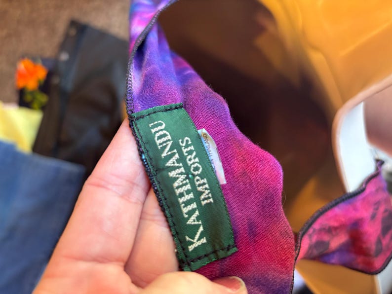 May include: A tie-dye garment featuring vibrant shades of purple, pink, and blue. A rectangular green fabric label is stitched onto the inner neckline, displaying the white text KATHMANDU IMPORTS in capital letters with a smaller tag underneath.