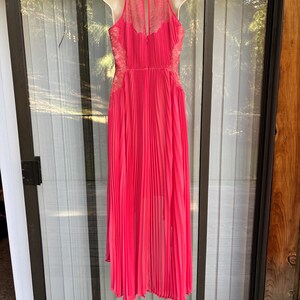 Puede incluir: Un vestido plisado rosa brillante con cuello halter y detalles de encaje. El vestido, exhibido en un maniqu&iacute;, presenta un dise&ntilde;o fluido y largo hasta el suelo. El vestido es de un tono rosa vibrante.