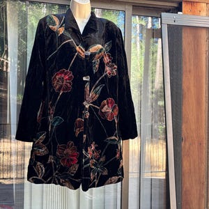 May include: A black velvet jacket featuring a long-sleeve design, a collared neckline, and a front button closure. The garment is decorated with an embroidered floral pattern in shades of red, green, and gold, extending across the front panels.