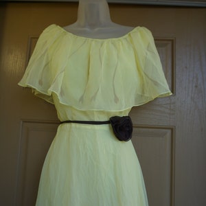 May include: A pale yellow dress with a flowing skirt and a ruffled overlay at the shoulders. The dress is cinched at the waist with a thin black belt and a dark brown floral accent. The dress is displayed on a mannequin.
