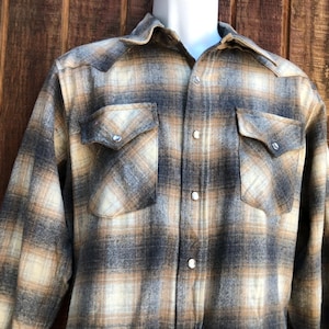 May include: A long-sleeved, button-down plaid shirt in shades of tan, gray, and cream. The shirt features two chest pockets with decorative flaps and silver snap closures. The collar and cuffs are visible.