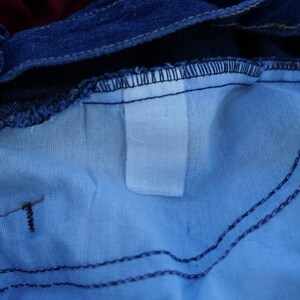 May include: Close-up of the inside of blue denim jeans. The image shows the waistband, the light blue lining, and the stitching details. A white label is visible.