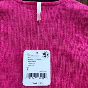 May include: A close-up of a raspberry sorbet colored top with a textured fabric. The top has a small white tag attached with the size 'S' indicated. The tag also includes a barcode and the price.