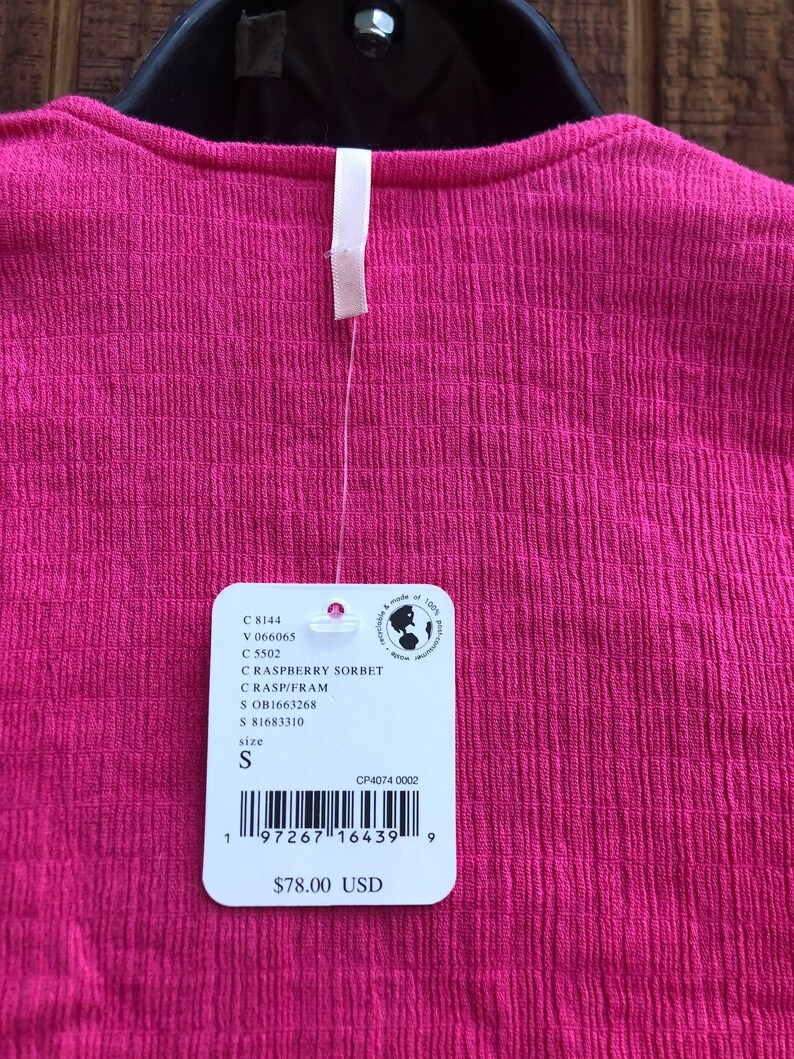 May include: A close-up of a raspberry sorbet colored top with a textured fabric. The top has a small white tag attached with the size 'S' indicated. The tag also includes a barcode and the price.