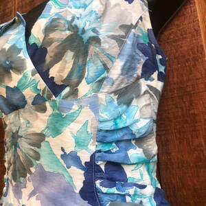 May include: A sleeveless dress with a V-neckline, featuring a floral pattern in shades of blue, teal, and gray on a white background. The fabric has a slightly sheer quality, and the dress has gathered detailing on the side.