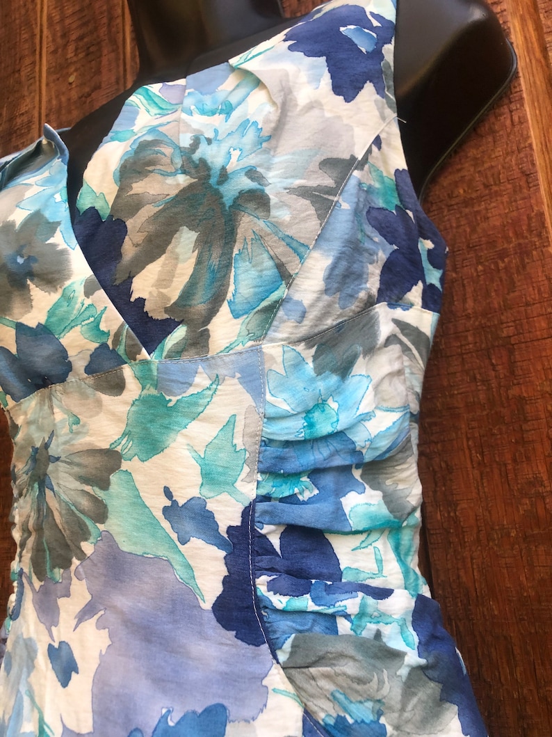 May include: A sleeveless dress with a V-neckline, featuring a floral pattern in shades of blue, teal, and gray on a white background. The fabric has a slightly sheer quality, and the dress has gathered detailing on the side.