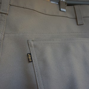 May include: Close-up of tan-colored pants with a back pocket and a small black tag with gold lettering. The pants are made of a textured fabric and have visible stitching details.