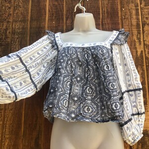 Puede incluir: Una blusa estampada en azul y blanco con mangas largas. Las mangas tienen un dise&ntilde;o de rayas florales y est&aacute;n adornadas con cinta azul oscuro. El corpi&ntilde;o presenta un patr&oacute;n geom&eacute;trico y un escote cuadrado. La blusa se exhibe en un maniqu&iacute;.