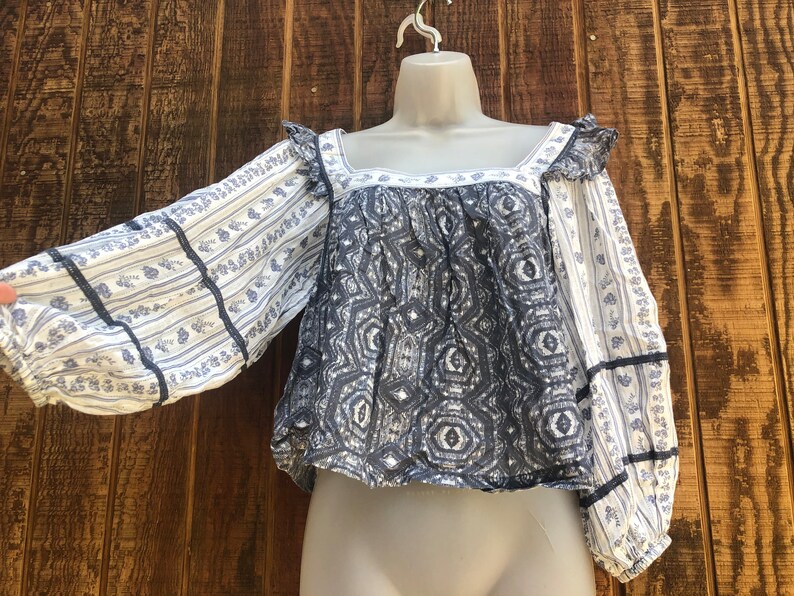 Puede incluir: Una blusa estampada en azul y blanco con mangas largas. Las mangas tienen un dise&ntilde;o de rayas florales y est&aacute;n adornadas con cinta azul oscuro. El corpi&ntilde;o presenta un patr&oacute;n geom&eacute;trico y un escote cuadrado. La blusa se exhibe en un maniqu&iacute;.