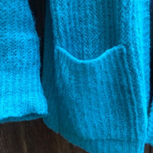 May include: A teal blue knitted cardigan with a pocket on the front.