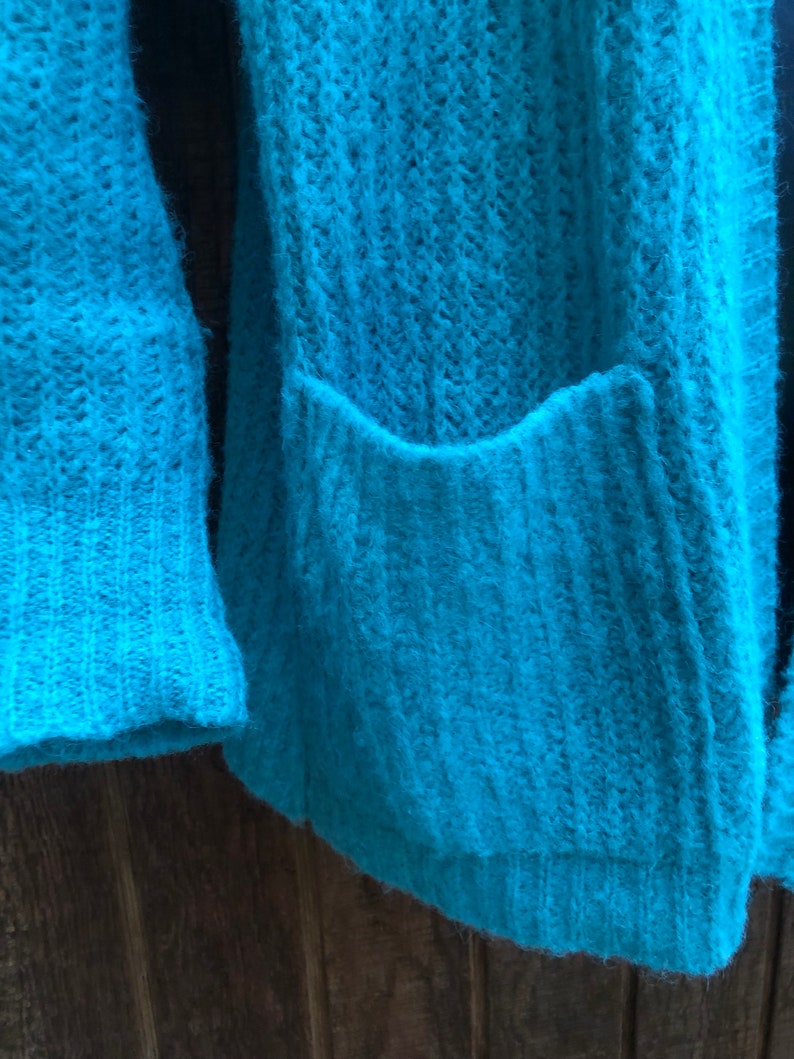 May include: A teal blue knitted cardigan with a pocket on the front.