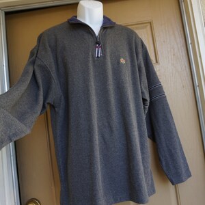 May include: A gray long-sleeve pullover shirt with a quarter-zip neckline and a small logo on the chest. The shirt has a navy blue collar and cuffs, with white and navy stripes on the sleeves.