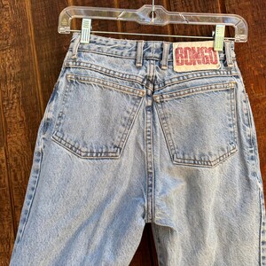 May include: Light blue denim jeans featuring a high-waisted design with two back pockets, a central zipper fly, and a rectangular white patch on the waistband with the red text BONGO printed in a bold, blocky font, suspended on a clear plastic hanger.