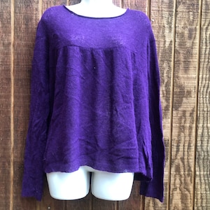 May include: A long-sleeved, purple sweater with a scoop neckline. The sweater has a slightly textured appearance and a relaxed fit. The background is a wooden wall.