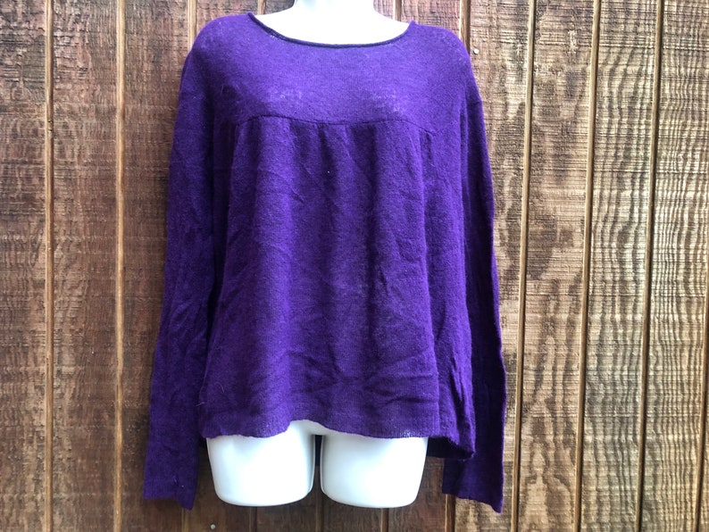 May include: A long-sleeved, purple sweater with a scoop neckline. The sweater has a slightly textured appearance and a relaxed fit. The background is a wooden wall.