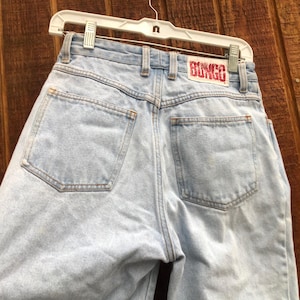 May include: Light blue denim jeans with a high waist, featuring two back pockets and a red "BONGO" label. The jeans are hanging on a white hanger against a wooden background. The jeans are a light wash.